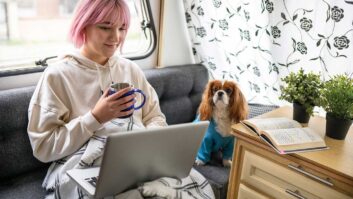 A woman on her laptop drinking tea with a dog on the sofa next to her in a caravan