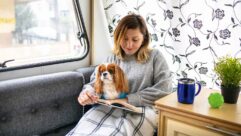 A smiling woman and a dog sitting under a blanket in a van as the woman reads a book